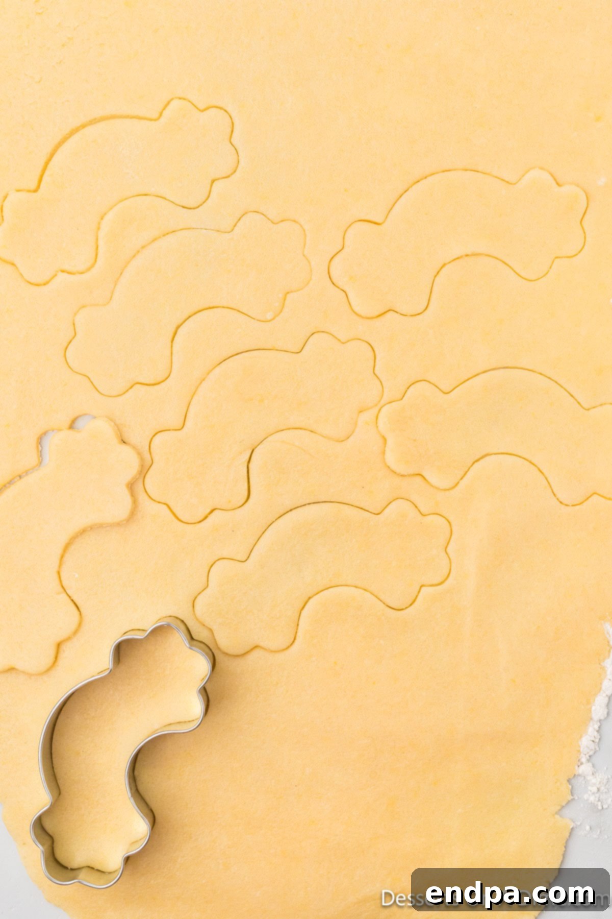 A rainbow-shaped cookie cutter pressed into rolled-out sugar cookie dough, ready for cutting.