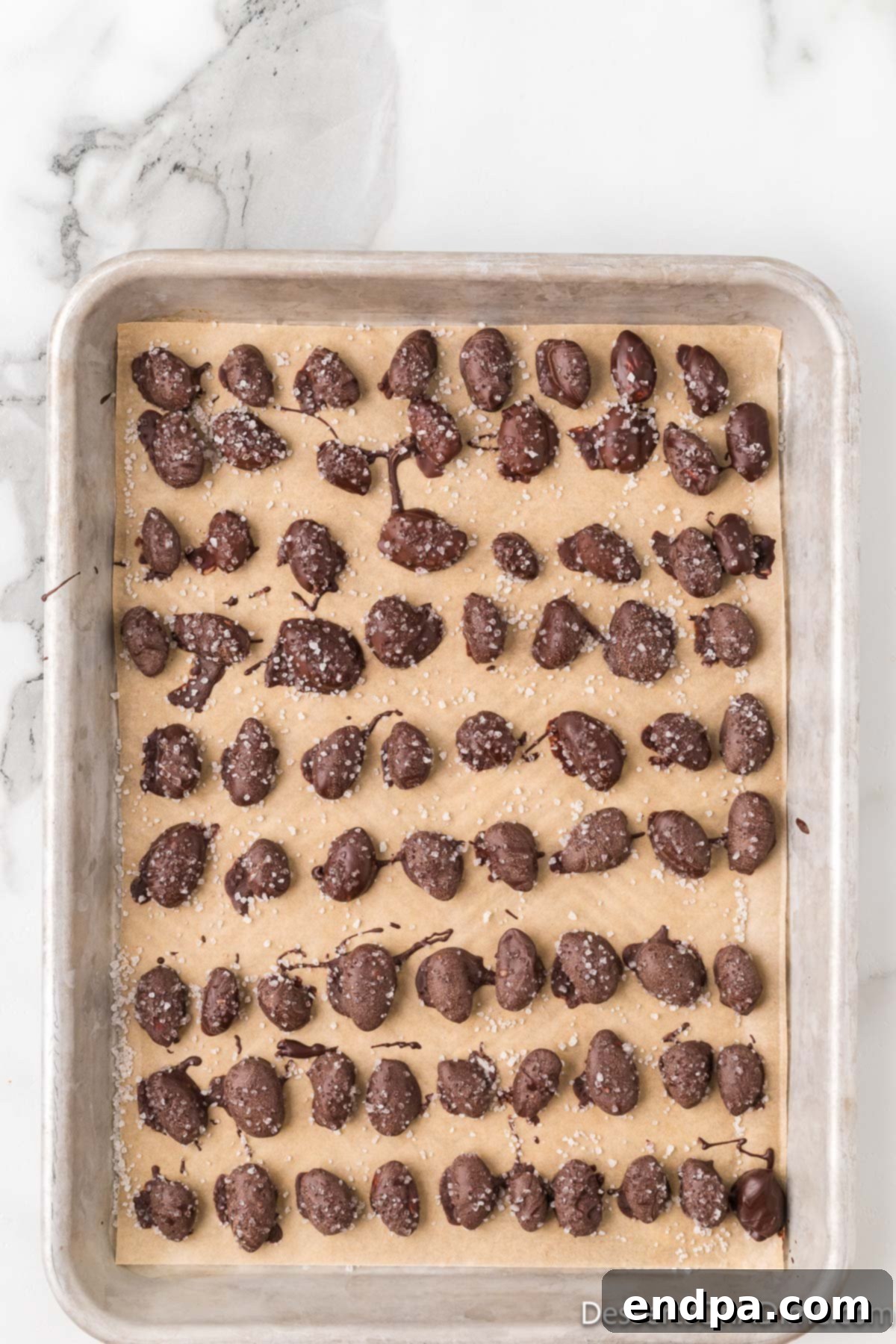 Decadent Dark Chocolate Almonds 8 Finished dark chocolate covered almonds, sprinkled with flaky sea salt, setting on a parchment-lined baking sheet.